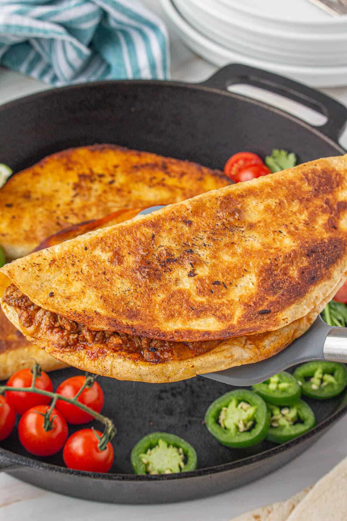 Crispy ground beef quesadillas in a cast iron skillet, with a spatula removing one of the cheesy quesadillas.