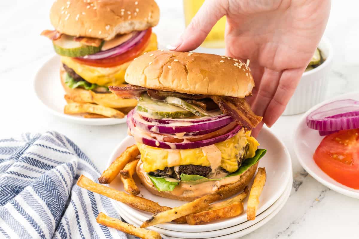 Steakhouse cheeseburgers on plates with crispy french fries. There's a hand reaching in to grab a burger.