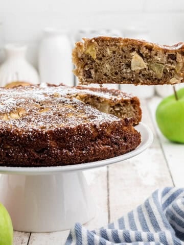 Sliced apple tea cake on a cake plate. A spatula is serving up a slice.