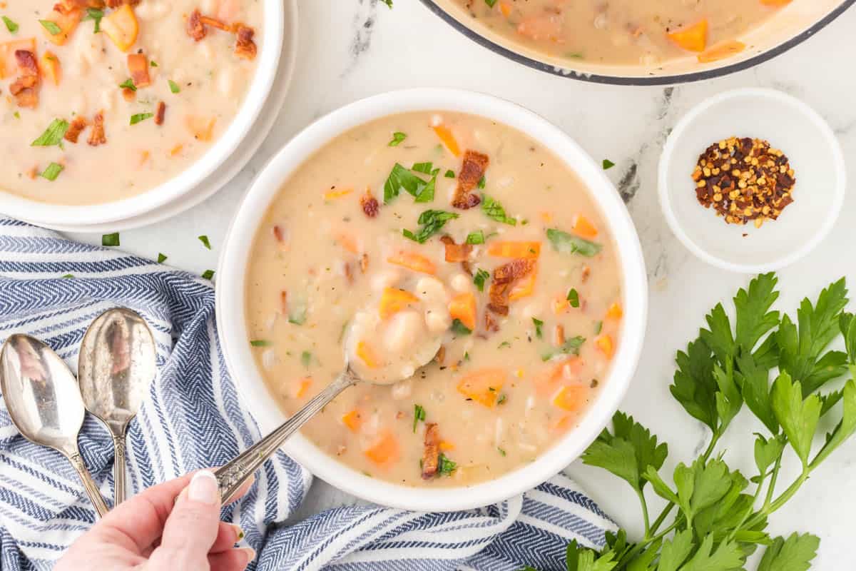 Bowls filled with bacon bean soup. There a hand with a spoon grabbing a bite of soup.