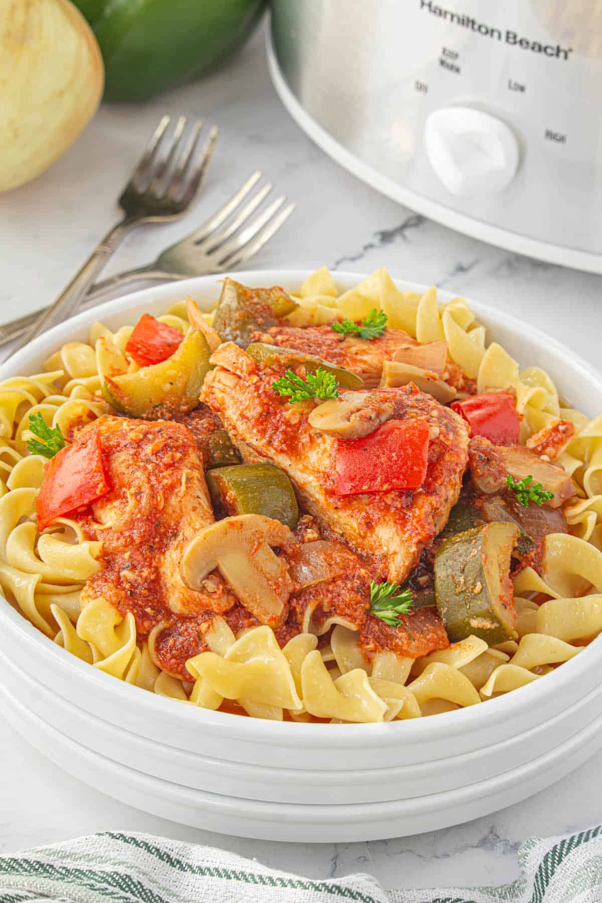 Saucy chicken cacciatore over noodles on a plate with a crockpot in the background.