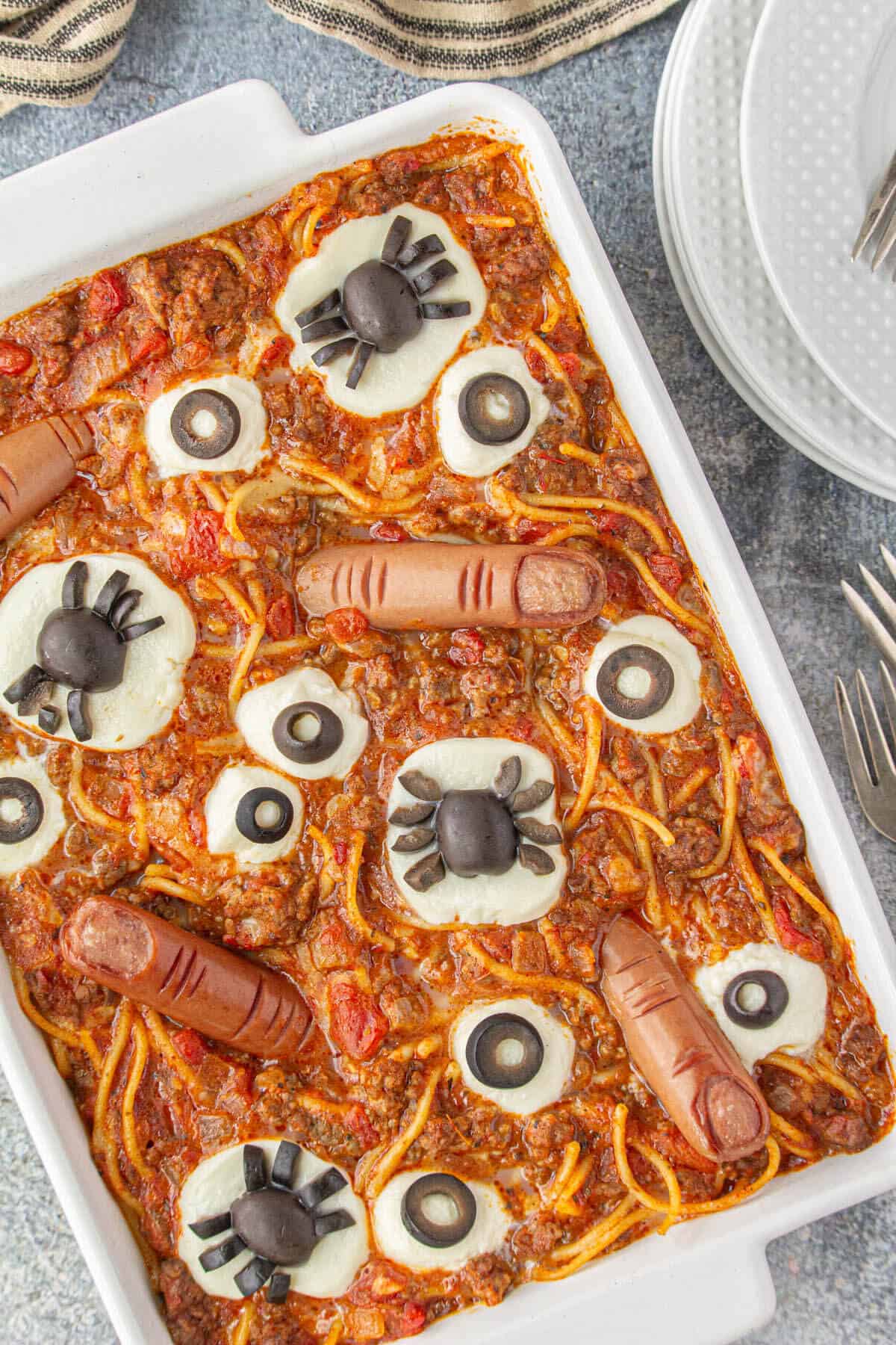 A casserole dish filled with creepy Halloween pasta recipe. It's topped with cheesy eyes, hot dog fingers, and spiders made from olives.