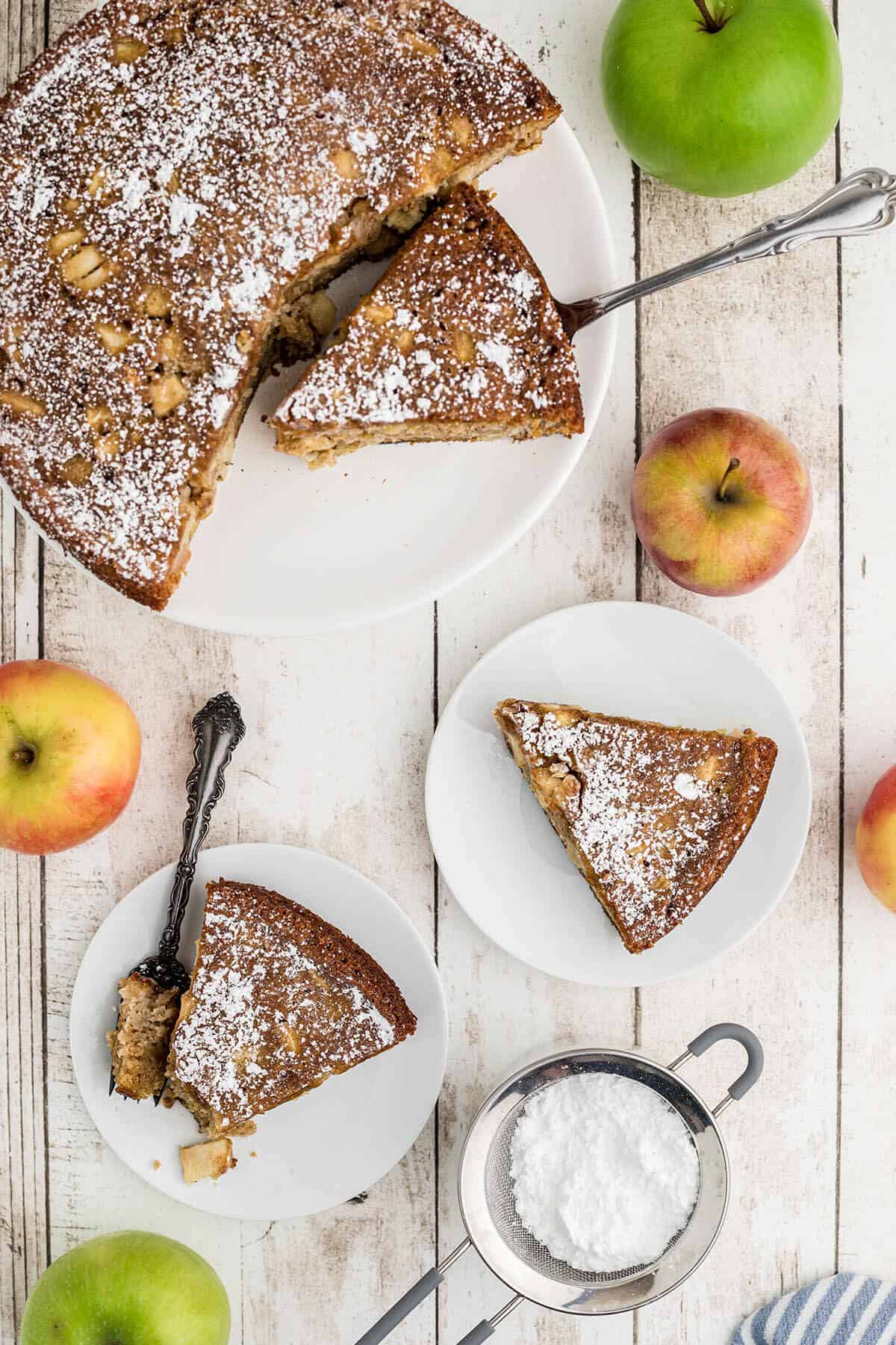 Sliced apple cake on serving plates with forks. The cake has been topped with powdered sugar.