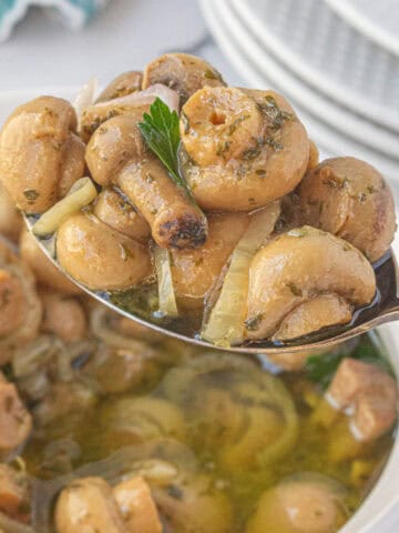 A spoon taking a scoop out of a bowl of pickled mushrooms.