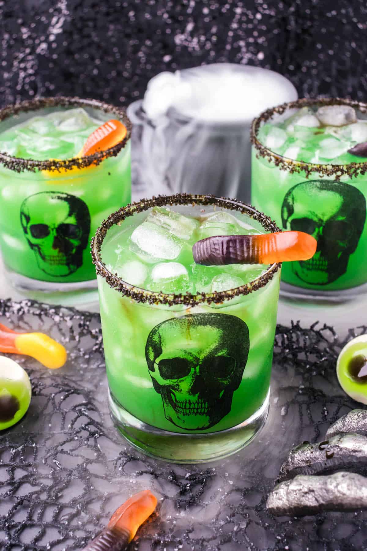 A trio of skull glasses fill with a witches brew cocktail. There's a bubbling cauldron in the background to add spooky fun to the photo.