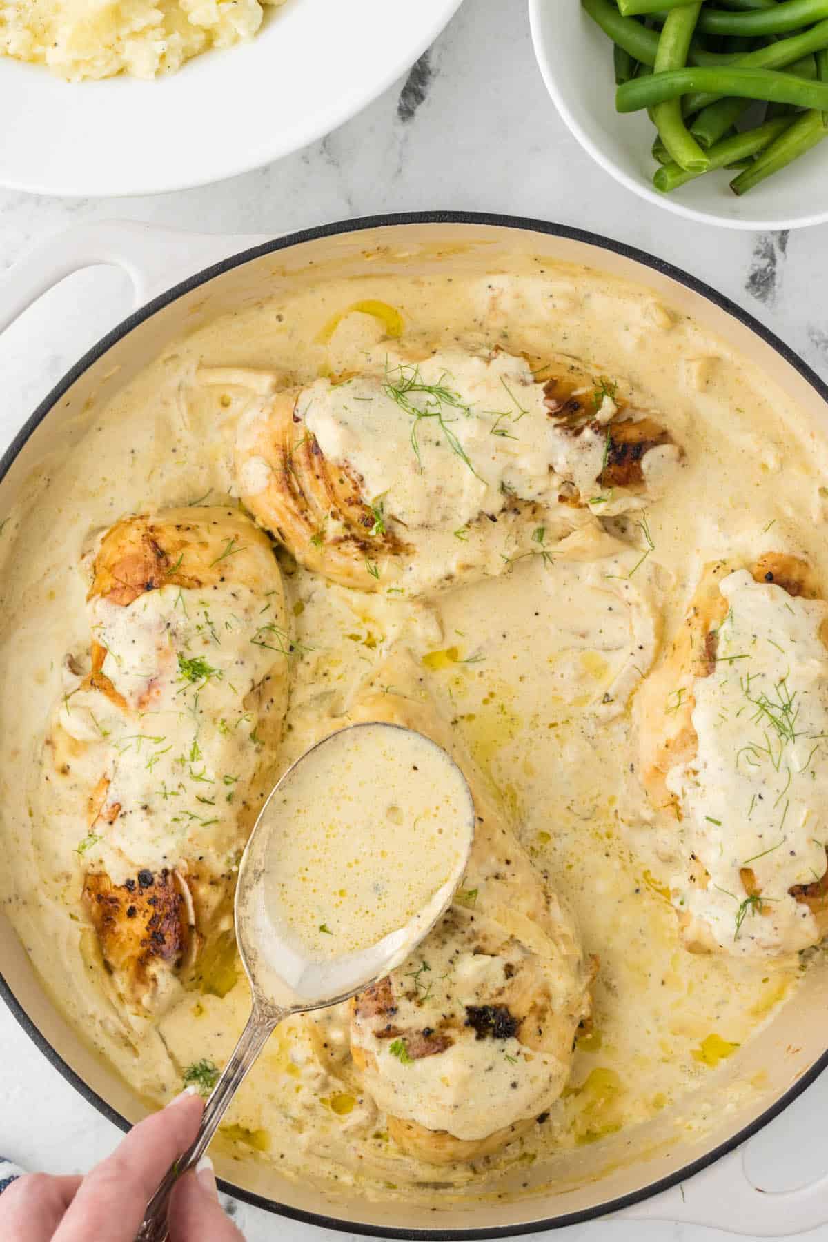 Baked chicken in a skillet with a spoon, scooping gravy over the chicken.