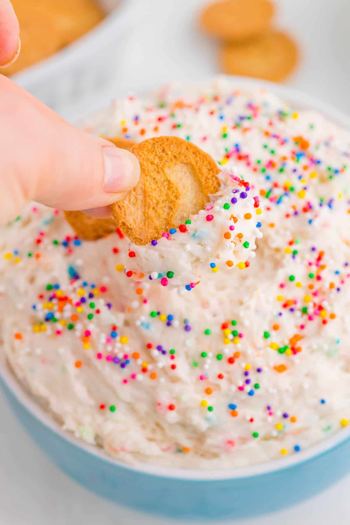 Funfetti Dunkaroo Dip in a blue bowl. A hand is dipping a vanilla wafer into the dip.