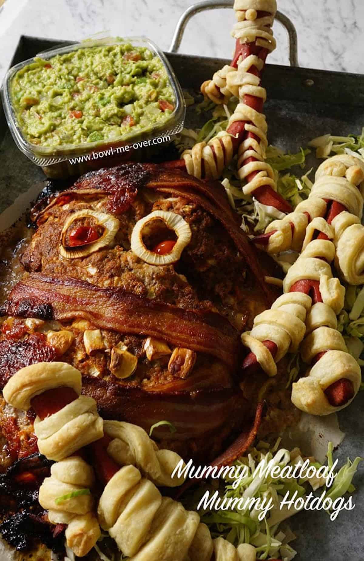 A Halloween meatloaf baked and ready to serve. It looks like a mummy and has been decorated with crispy bacon, garlic cloves for teeth and baby tomatoes for red eyes. With print overlay.