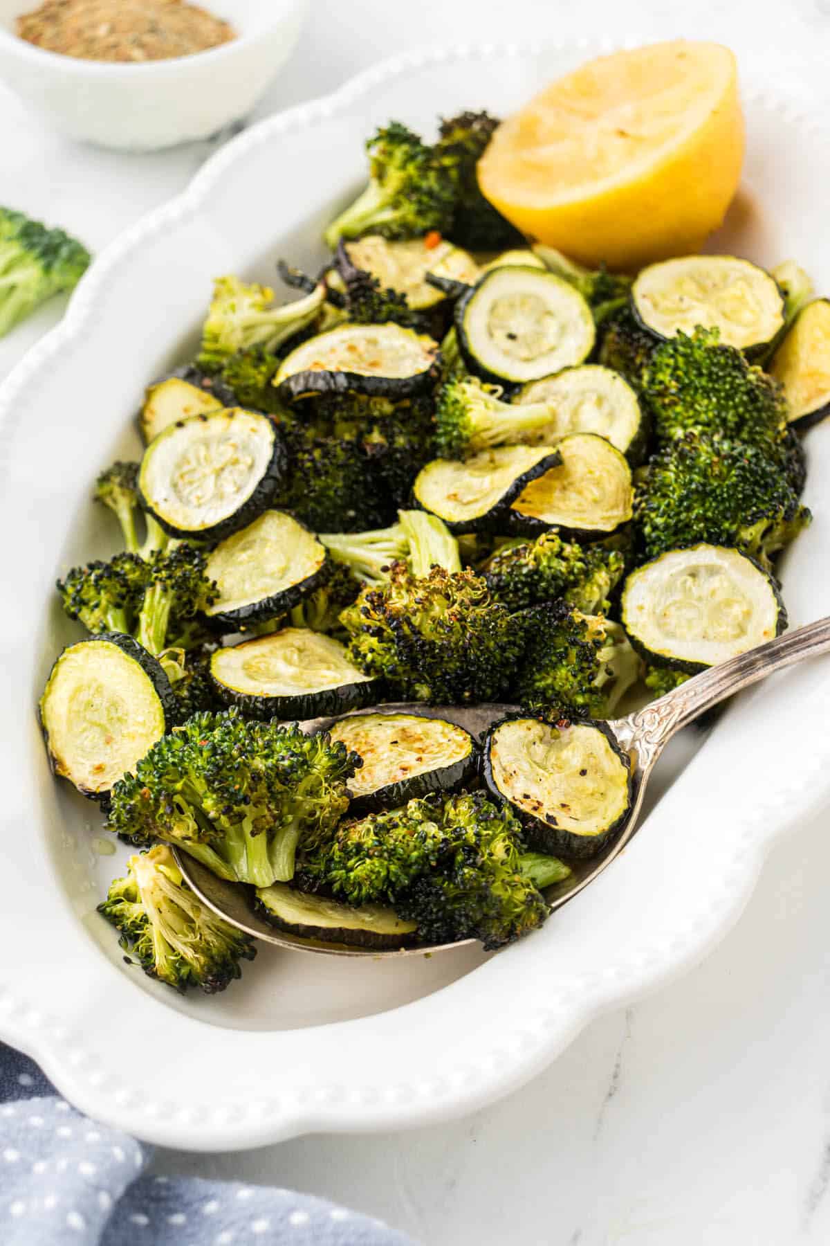 A pretty white platter filled with roasted broccoli and zucchini. Wtih a spoon for serving spoon.