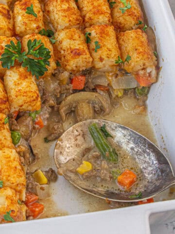 Tater tot shepherds pie casserole in a baking dish. A spoon has already removed a serving and it's laying in the casserole.