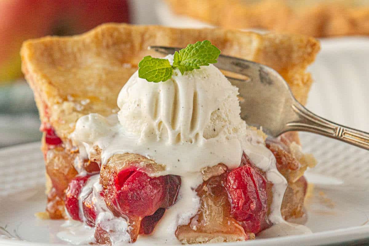 A slice of apple cranberry pie topped with a scoop of melted ice cream.