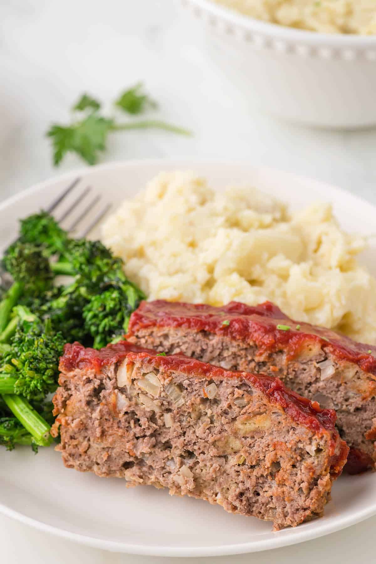 Slices of meatloaf on a plate, serve with steamed broccoli and mashed potatoes.