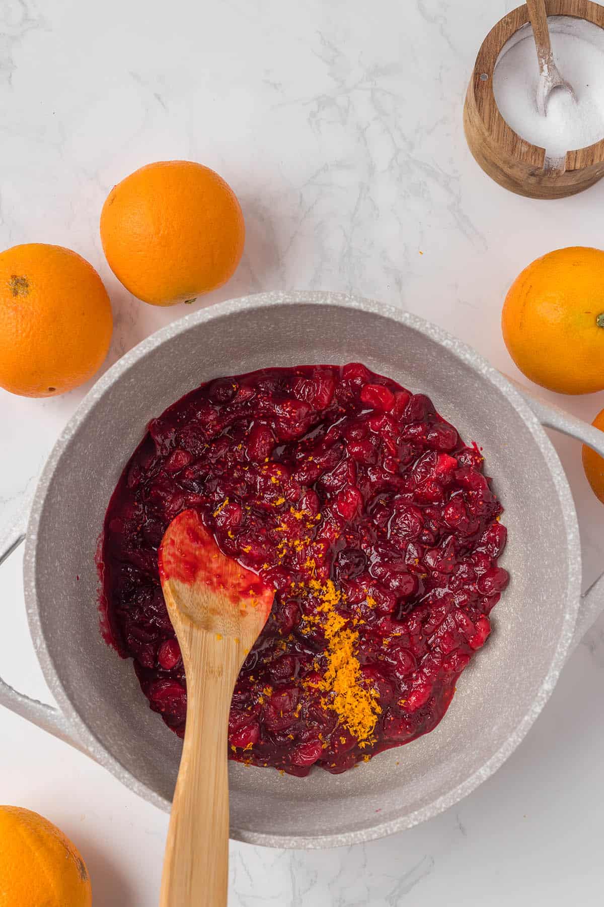 Cooked cranberries in a sauce pan with a wooden spoon stiring the sauce.