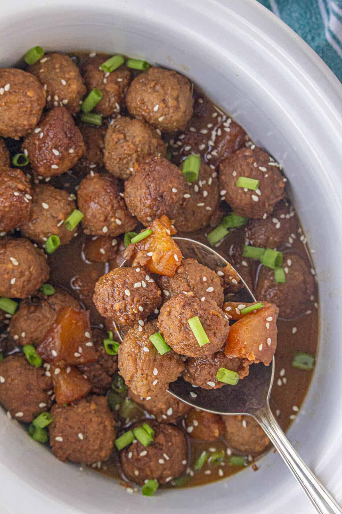 Teriyaki meatballs in a slow cooker, garnished with green onions and sesame seeds. With a serving spoon.