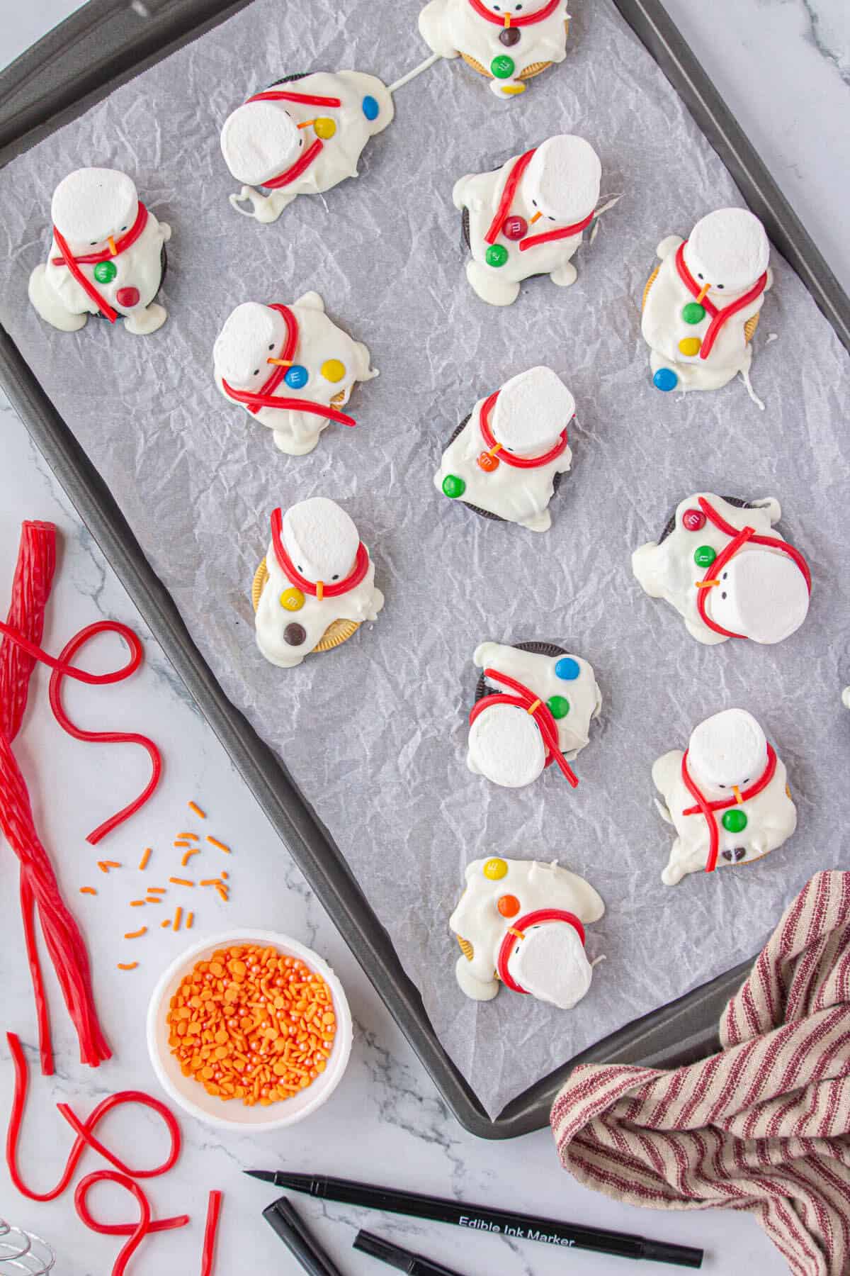 Melted snowmen cookies decorated with a sprinkle for a nose, eyes drawn on the marshmallow and a red licorice twizzle for a scarf.