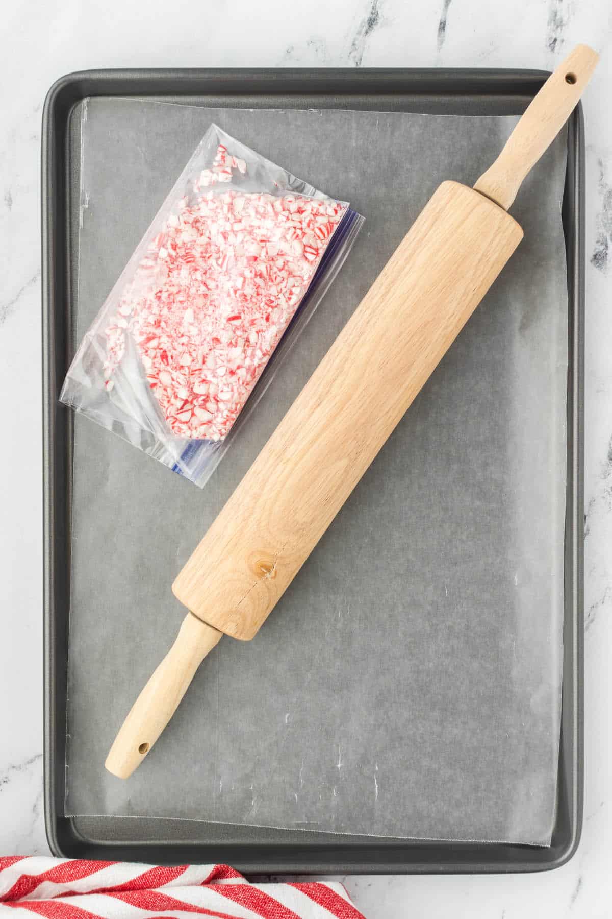 Crushed candy canes in a ziplock bag beside a rolling pin, on a cookie sheet.