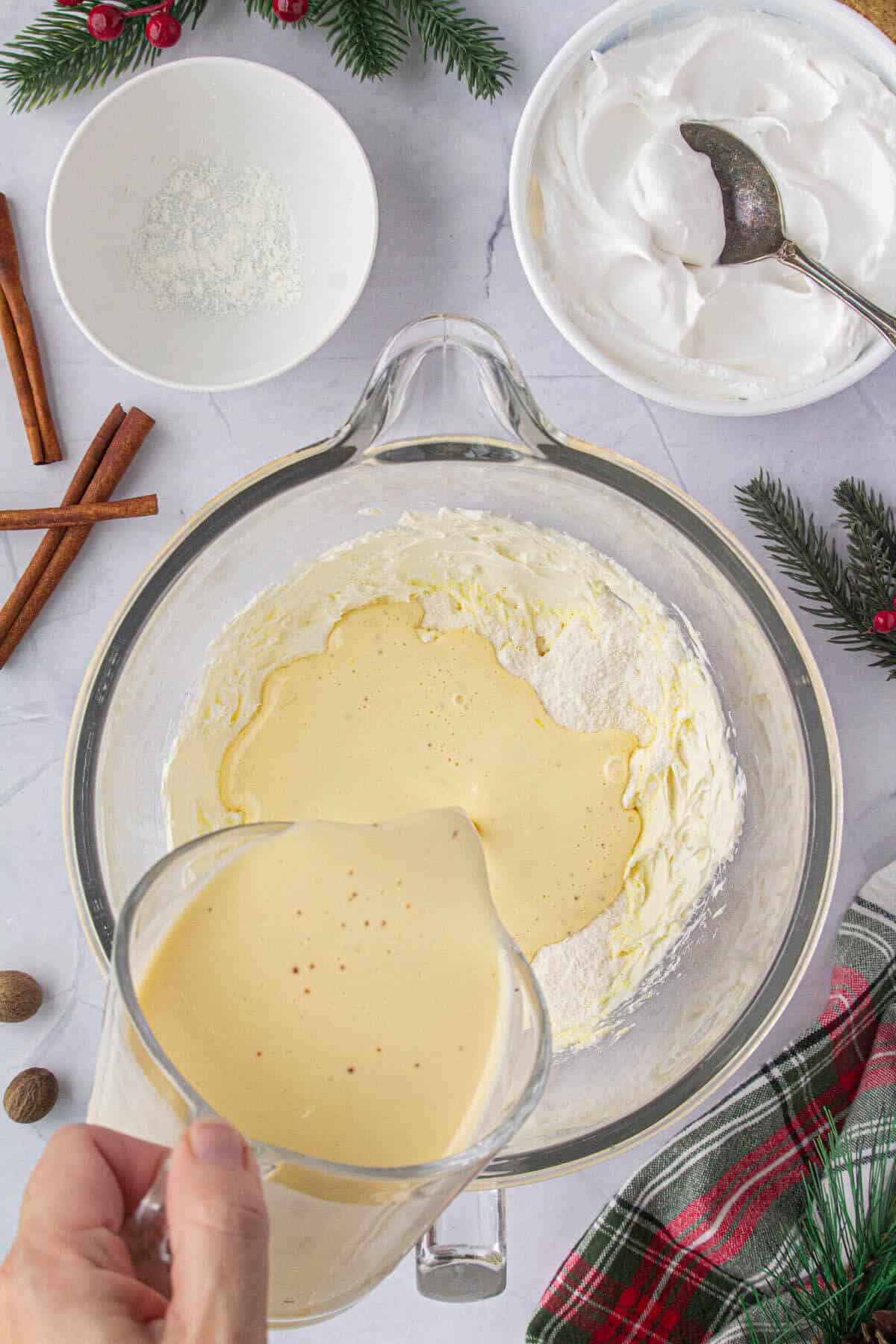 Egg nog being added to whipped cream cheese in a mixing bowl.