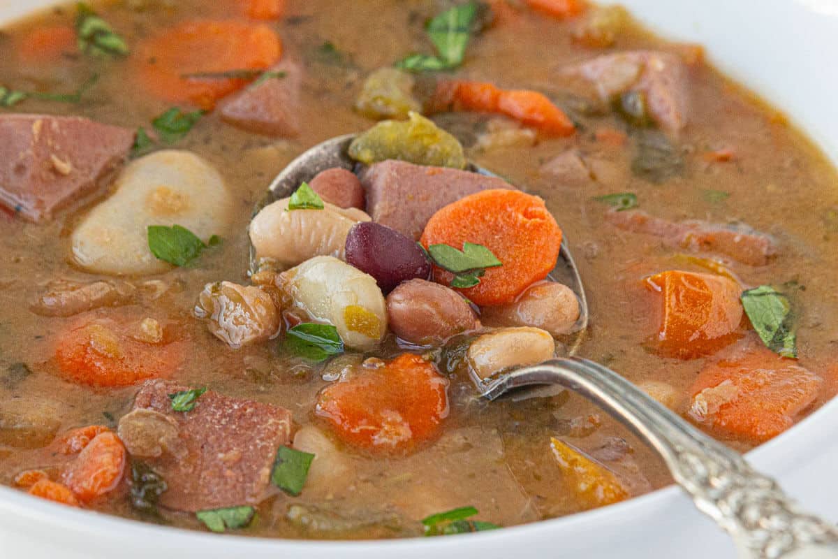 Bean soup filled with vegetables in a white bowl with a serving spoon.