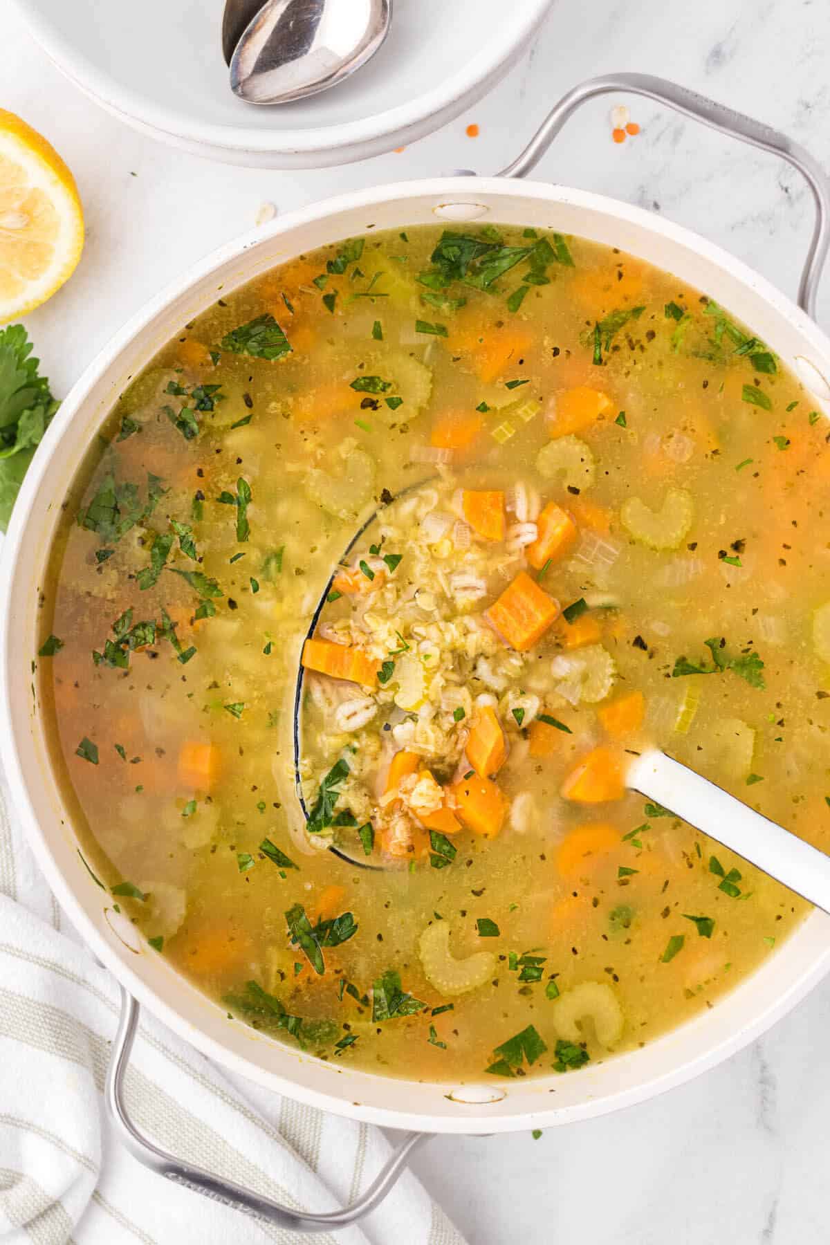 A large soup pot filled with a light barley and lentil soup. With a ladle to scoop out soup to serve.