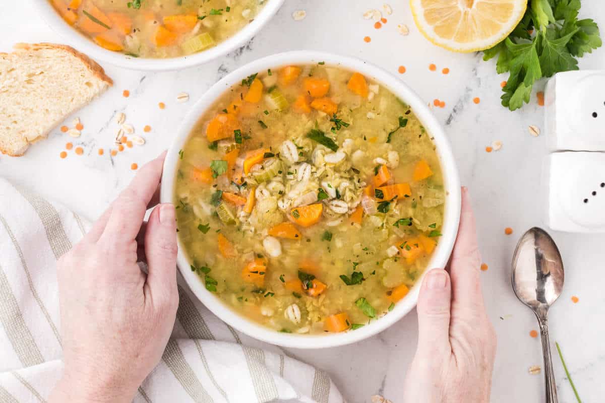 Barley and Lentil Soup in a bowl with hands reaching out and graphing the bowl of soup.