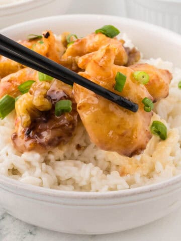 Juicy Asian shrimp in a bowl over rice. With a pair of chopsticks holding a piece of shrimp.