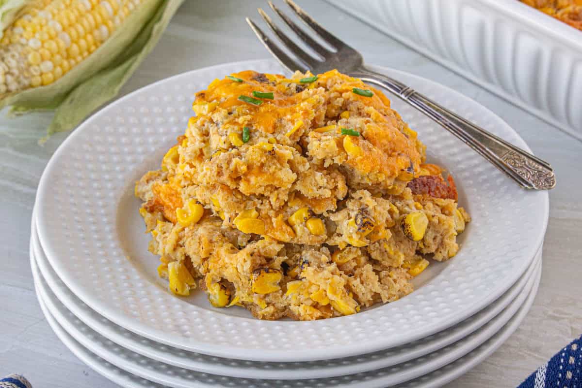 A stack of white plates. The top one is filled with Corn Casserole. A fork is off to the side.