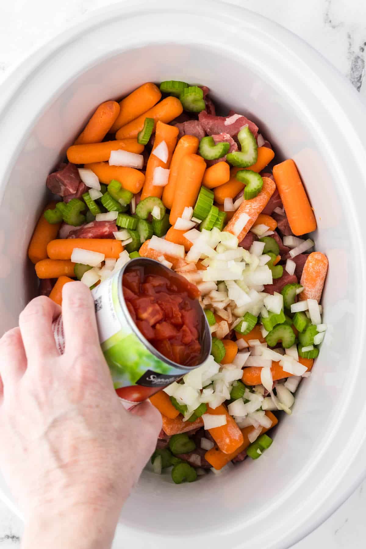 Chopped vegetables and beef chunks in a slow cooker, with a can of chopped tomatoes being added to the top.