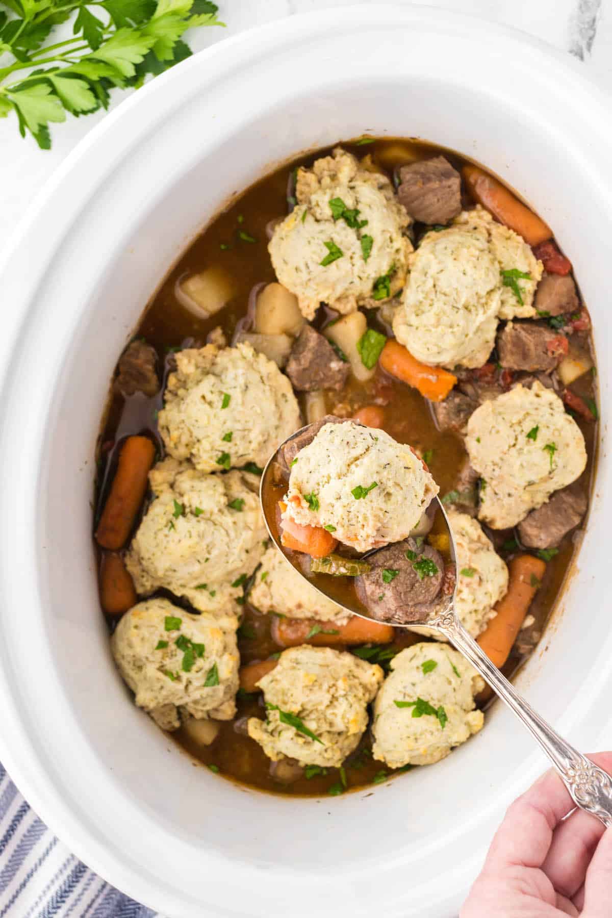 Beef stew in a crockpot topped with fluffy dumplings. With a serving spoon dishing out a serving.