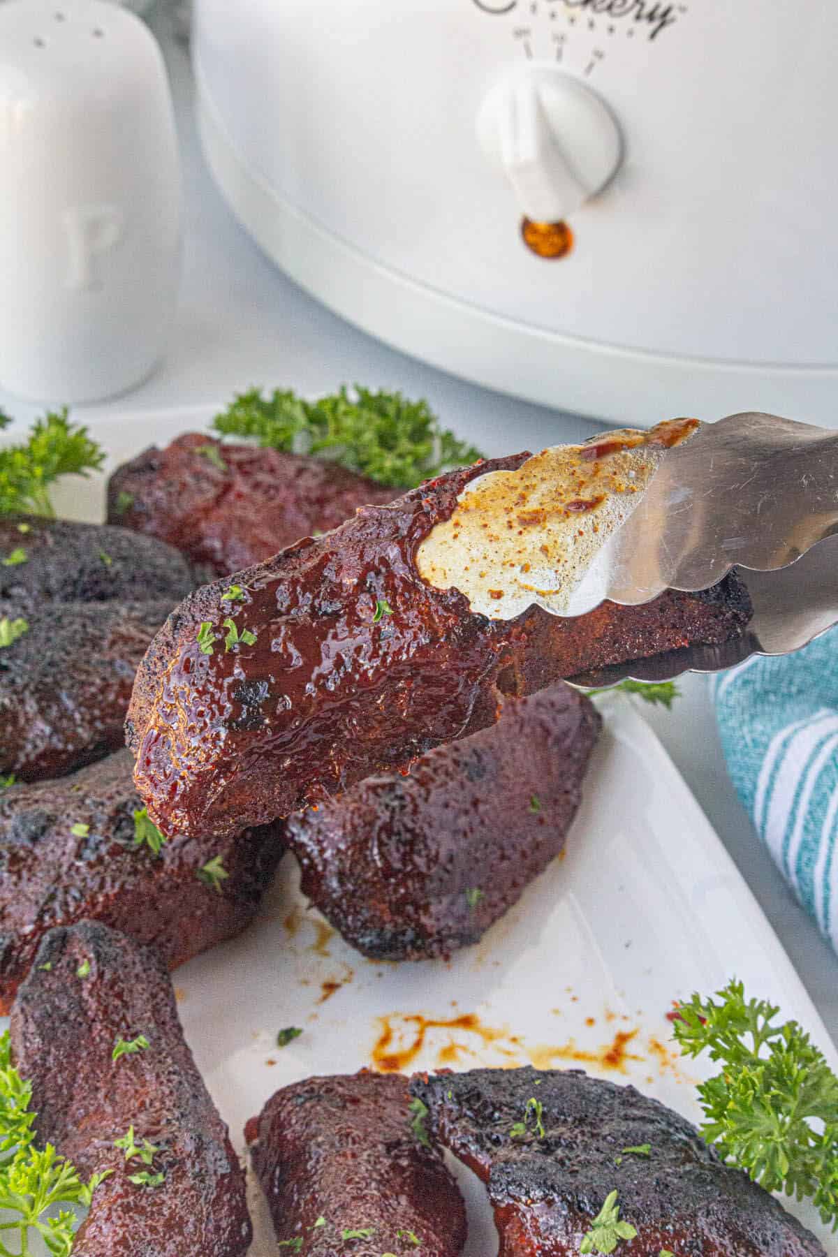 Slow Cooker Country Style Pork Ribs on a platter, with a set of tongs holding one of the ribs.