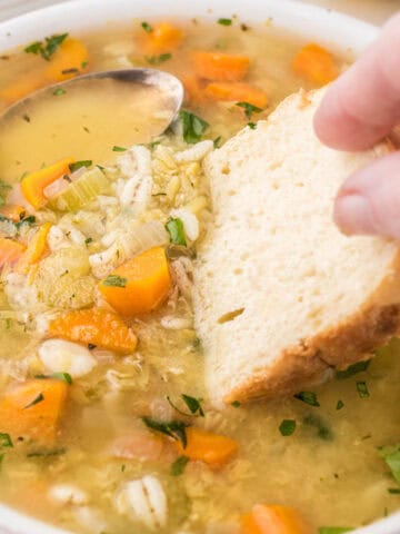 A bowl filled with lentil barley soup with a hunk of bread dunking into the broth.