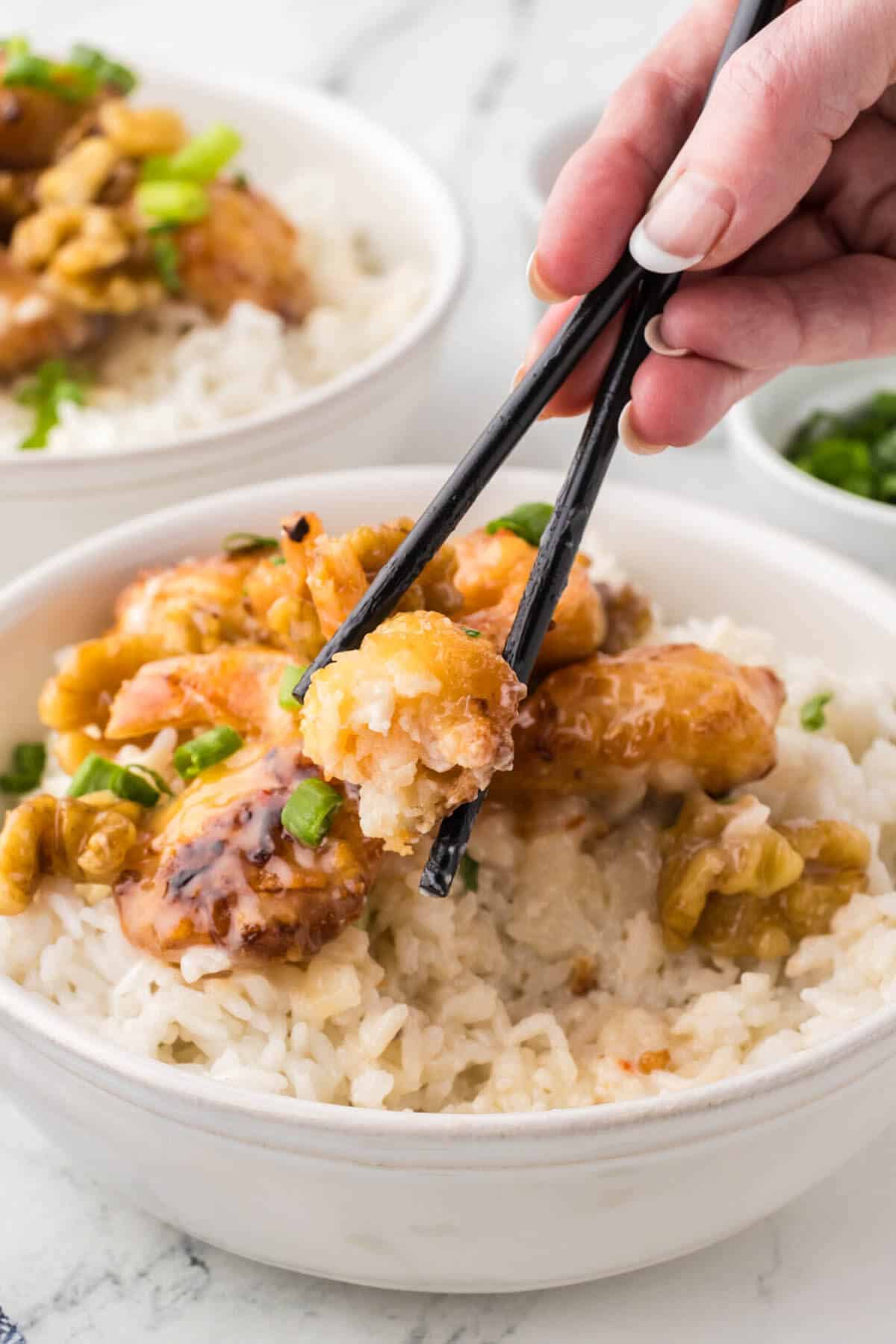 A bowl of rice topped with Panda Express Honey Walnut Shrimp. With a pair of chopsticks holding a shrimp.