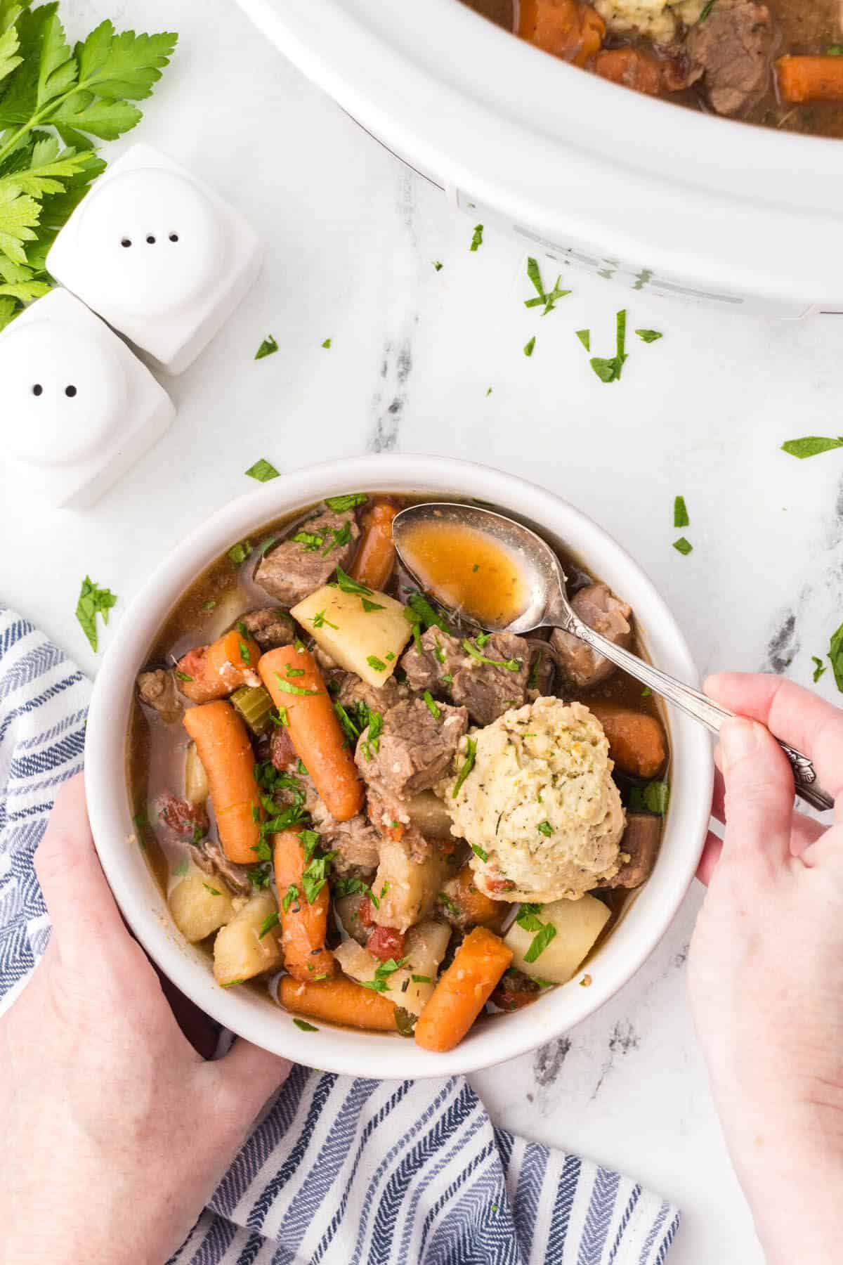 Slow cooker beef stew with dumplings in a bowl. Hands are wrapped around the bowl and dipping a spoon in to the soup to take a bite.