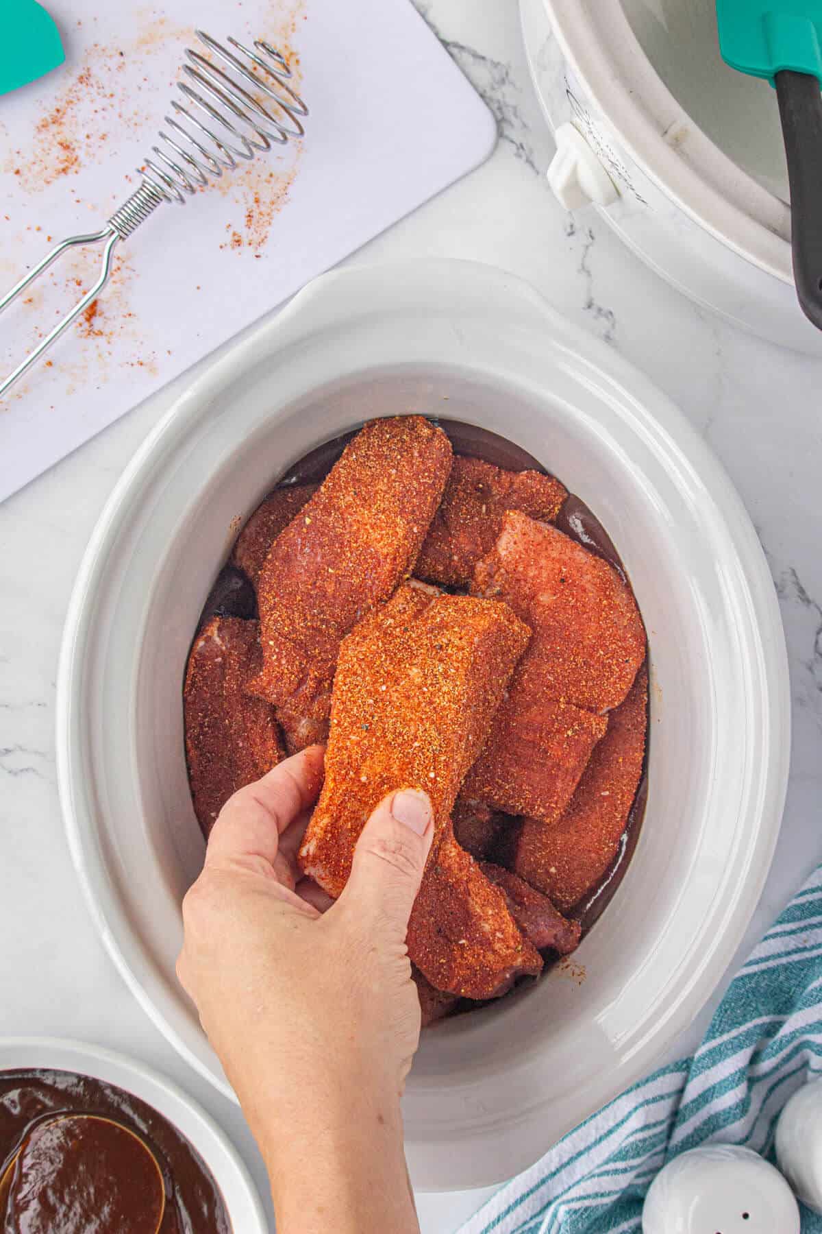 Seasoned ribs being added to a crockpot with bbq sauce.