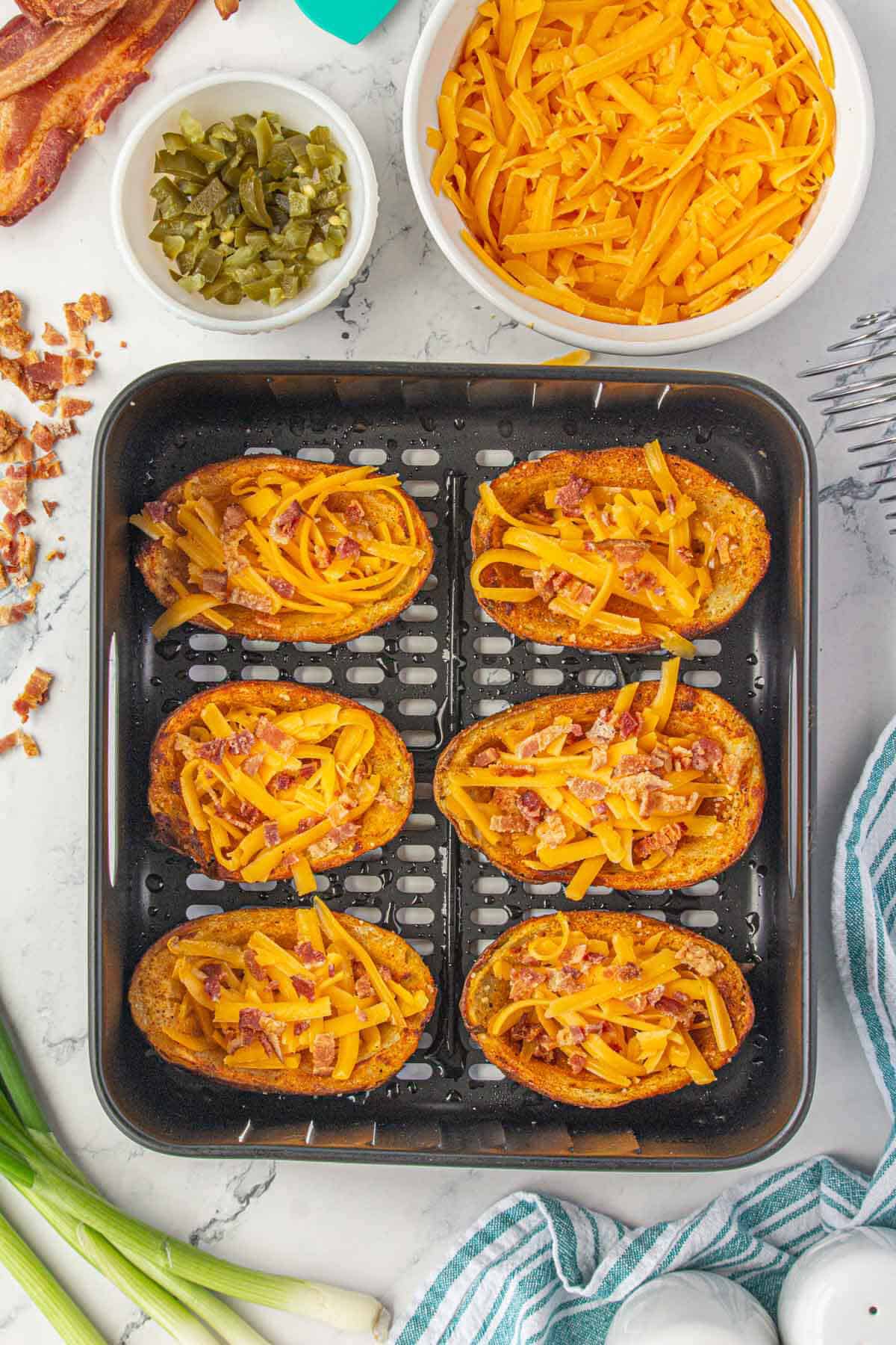 Crispy potato skins in an air fryer basket topped with shredded cheese and bacon bits.