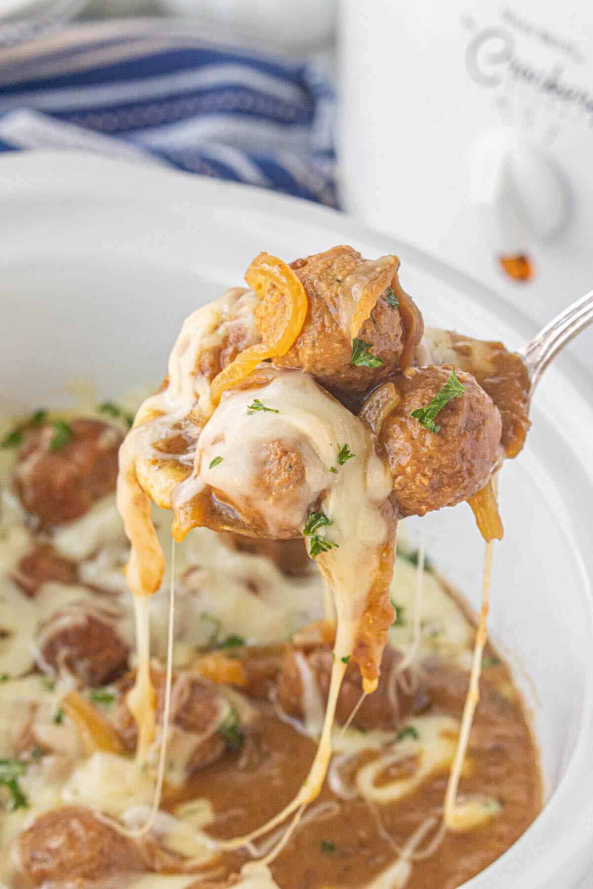 French onion meatballs in a crockpot with a serving spoon taking a big scoop. The cheese topping the meatballs is melty and looks luscious.