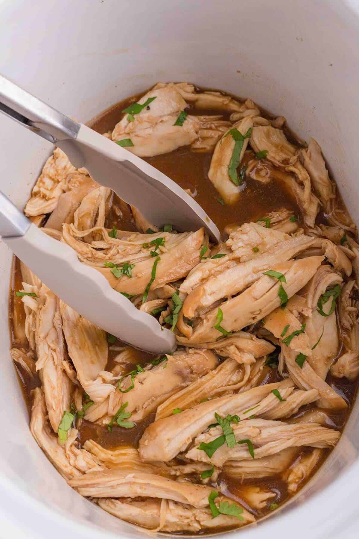 Shredded crockpot honey garlic chicken in a slow cooker, with serving tongs.