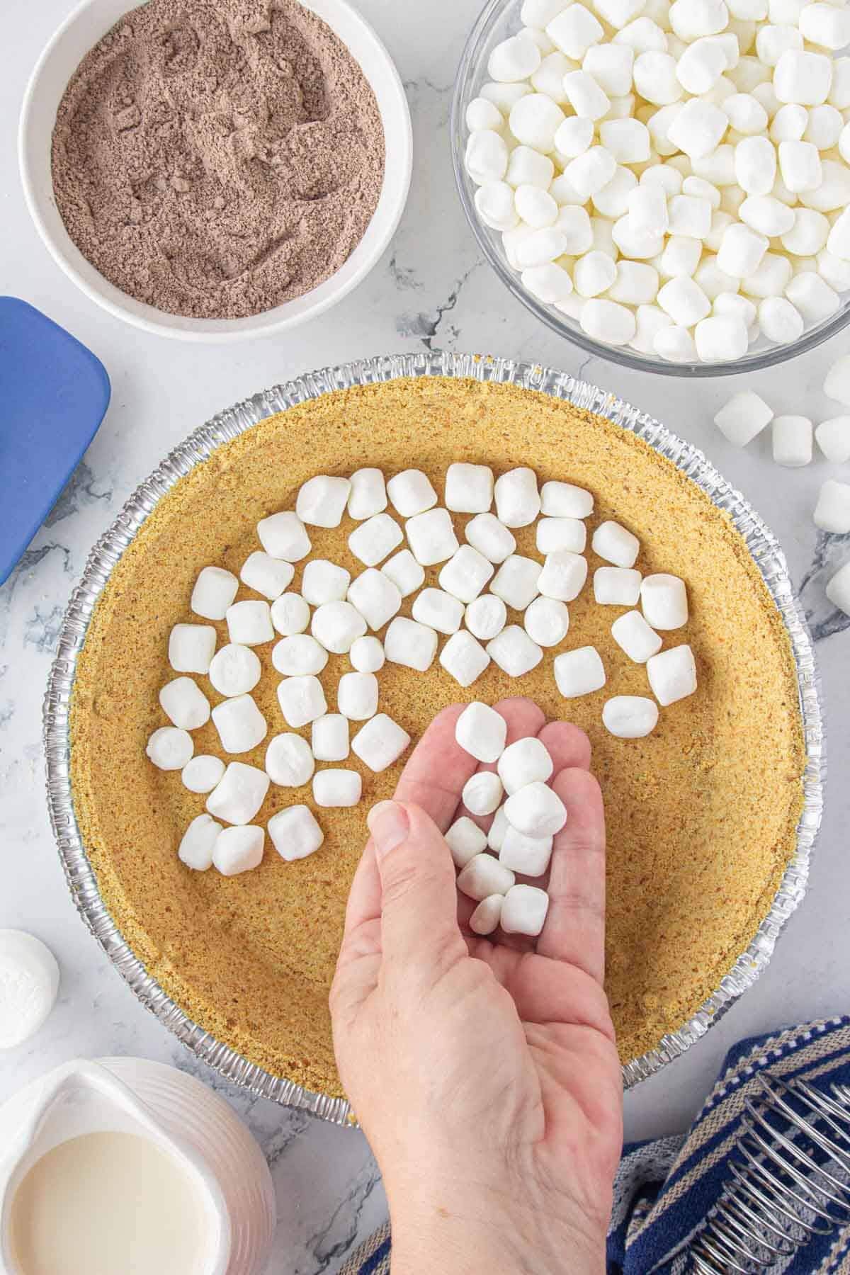 A handful of marshmallows being spread on the bottom of a graham cracker crust.