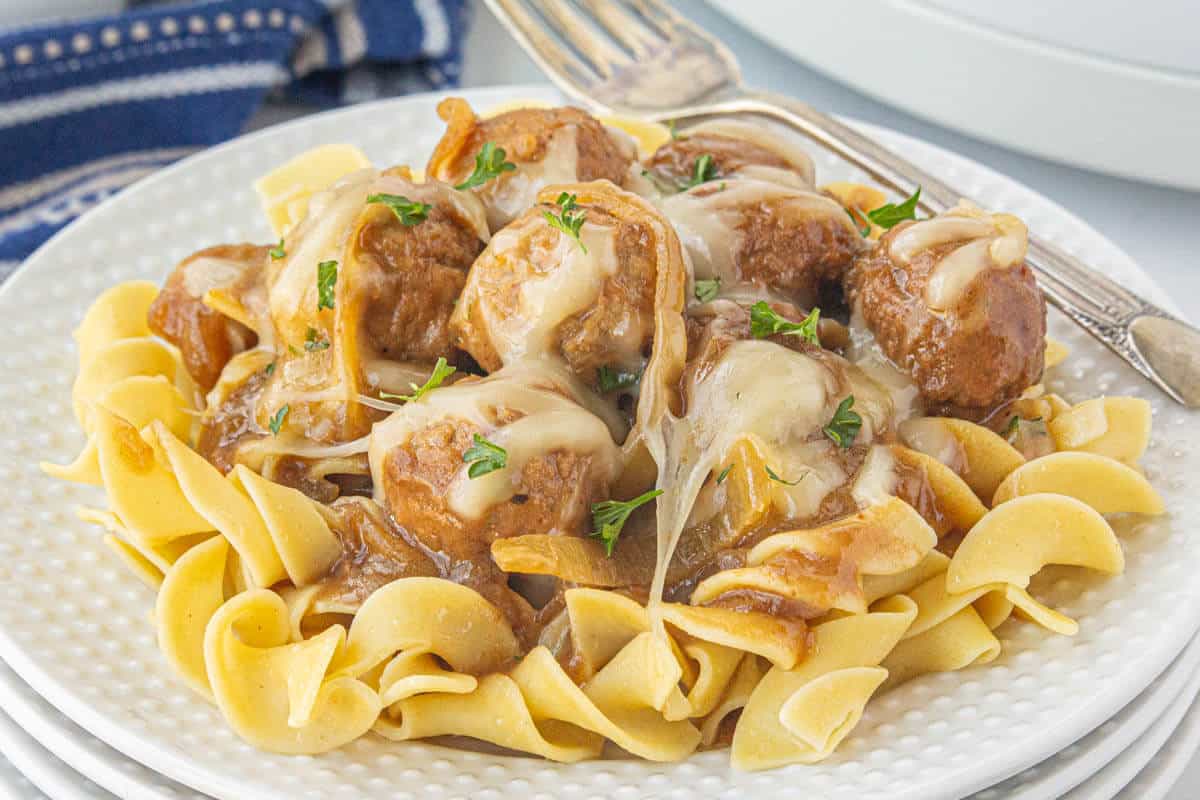 A plate of egg noodles topped with crockpot French onion meatballs. With a fork on the side of the plate.