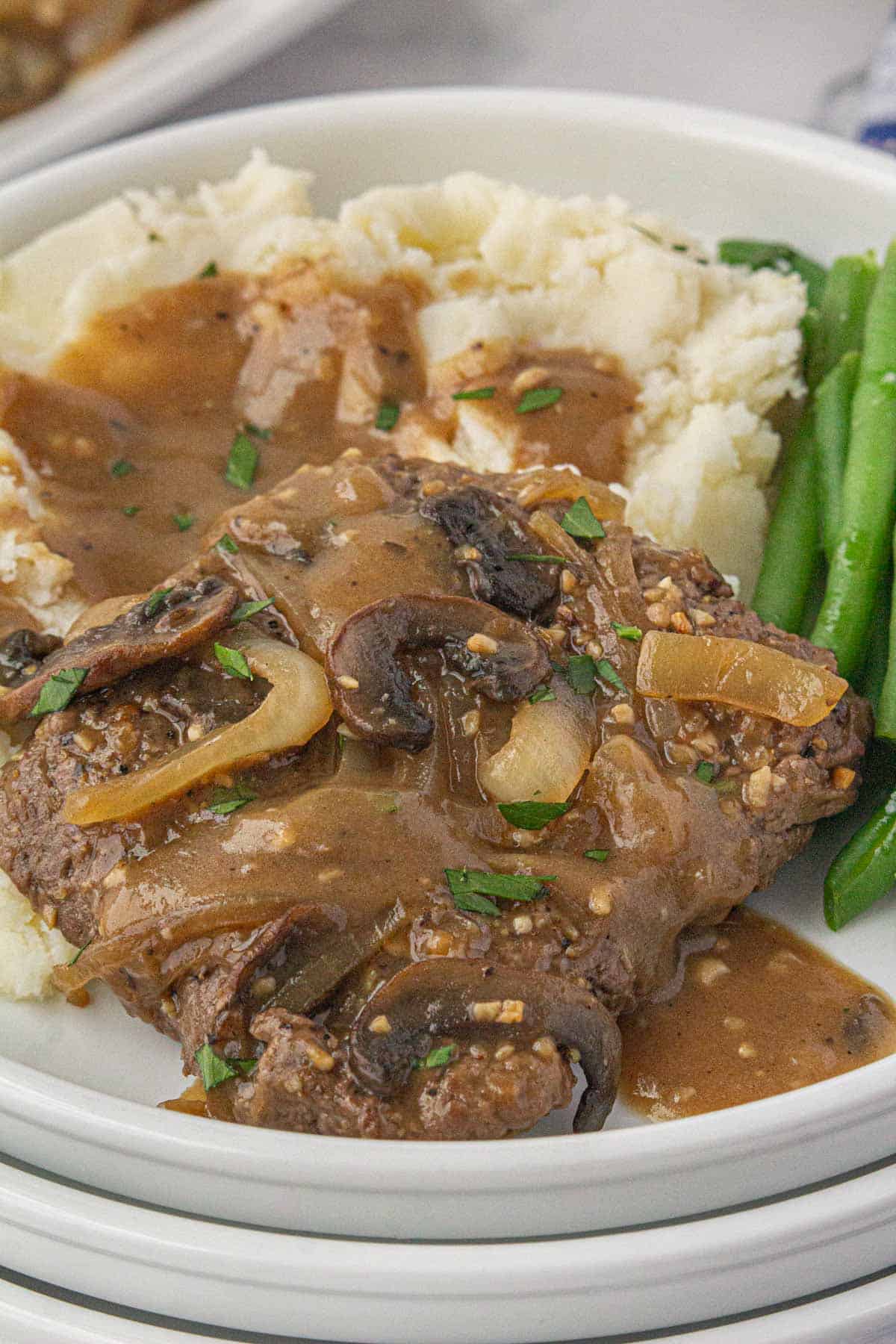 Smothered steak, potatoes and green beans on a plate. The mashed potatoes and steak is covered in gravy.