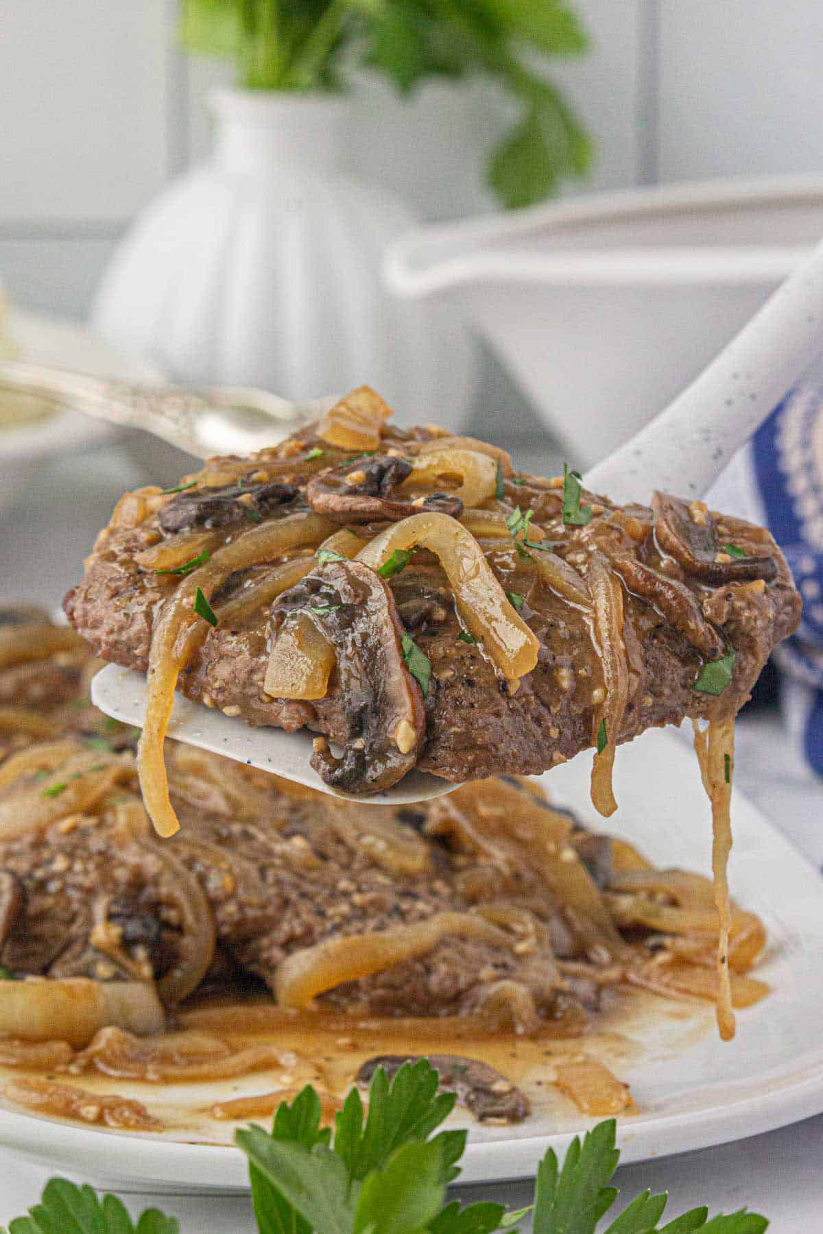 A platter of cooked cube steak smothered in an onion, mushroom gravy. With a spatula holding one of the steaks.