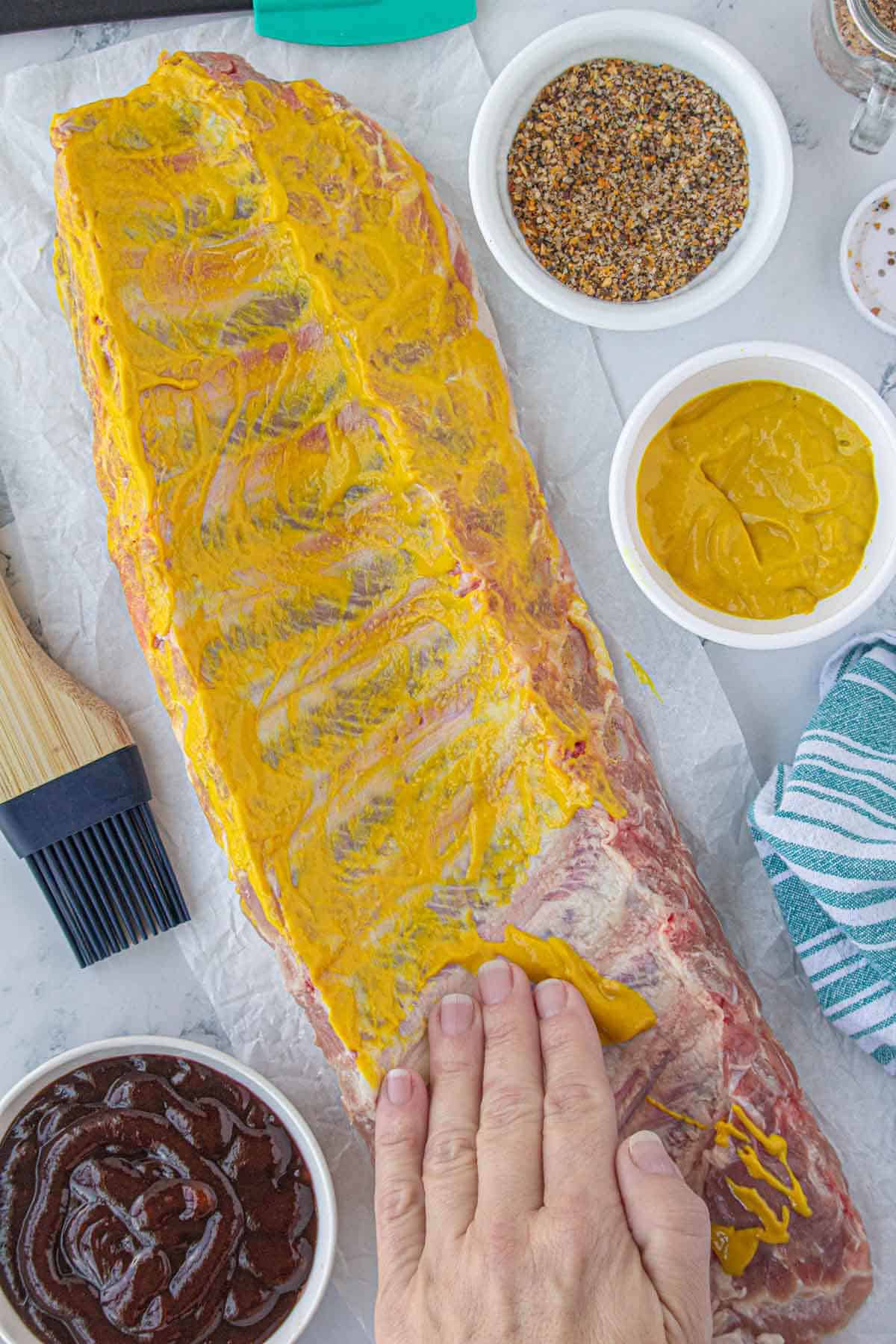 A hand slathering yellow mustard on a rack of pork spareribs.