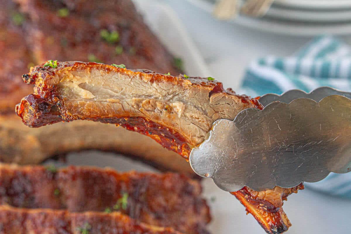 A pair of tongs holding a bbq pork rib that has been slowly cooked in the oven.