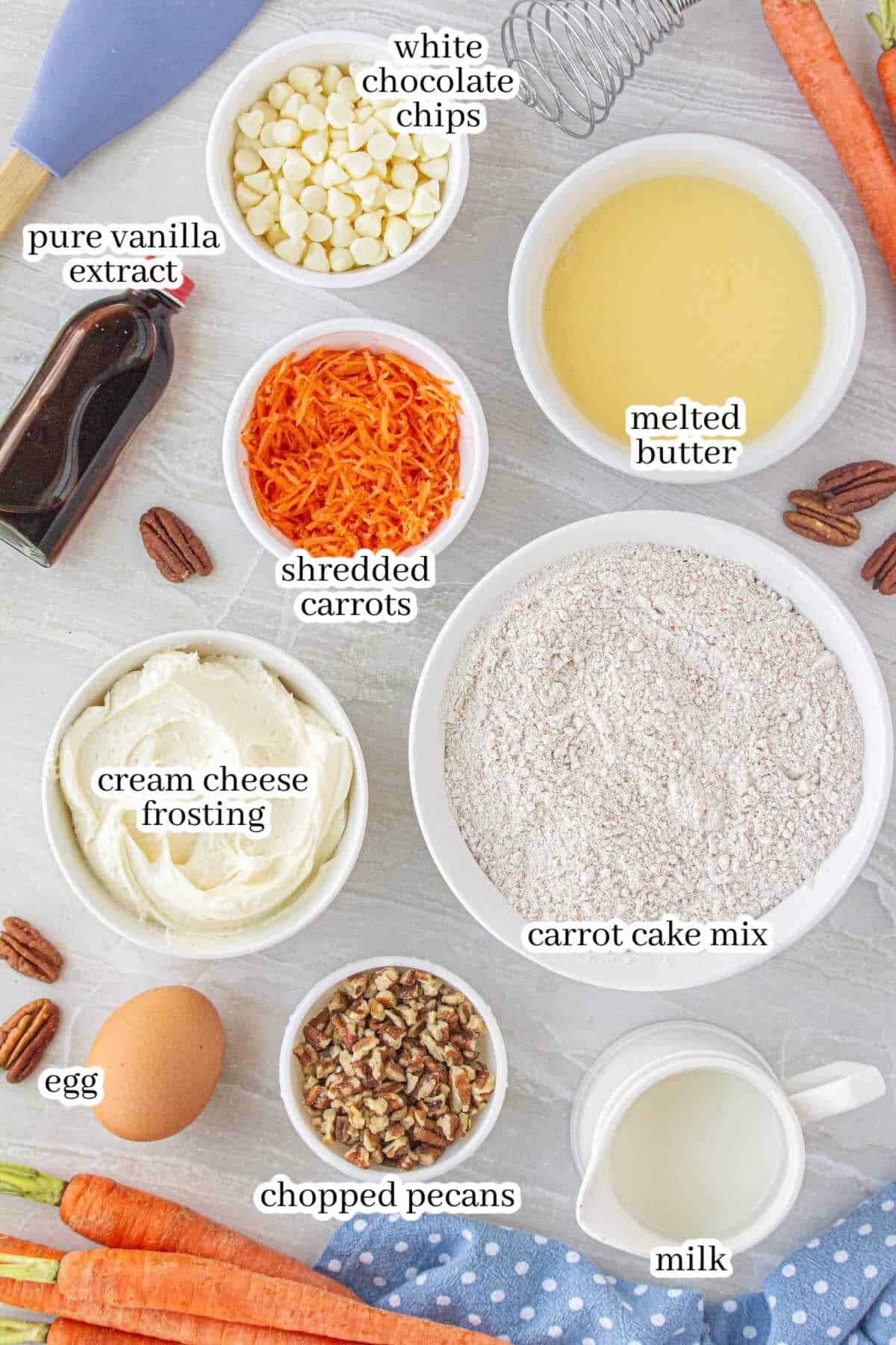 Overhead view of labeled ingredients for carrot cake blondies, including white chocolate chips, pure vanilla extract, shredded carrots, melted butter, spice cake mix, cream cheese frosting, an egg, chopped pecans, and milk, all arranged on a light countertop with whole carrots and a polka dot towel for decoration.