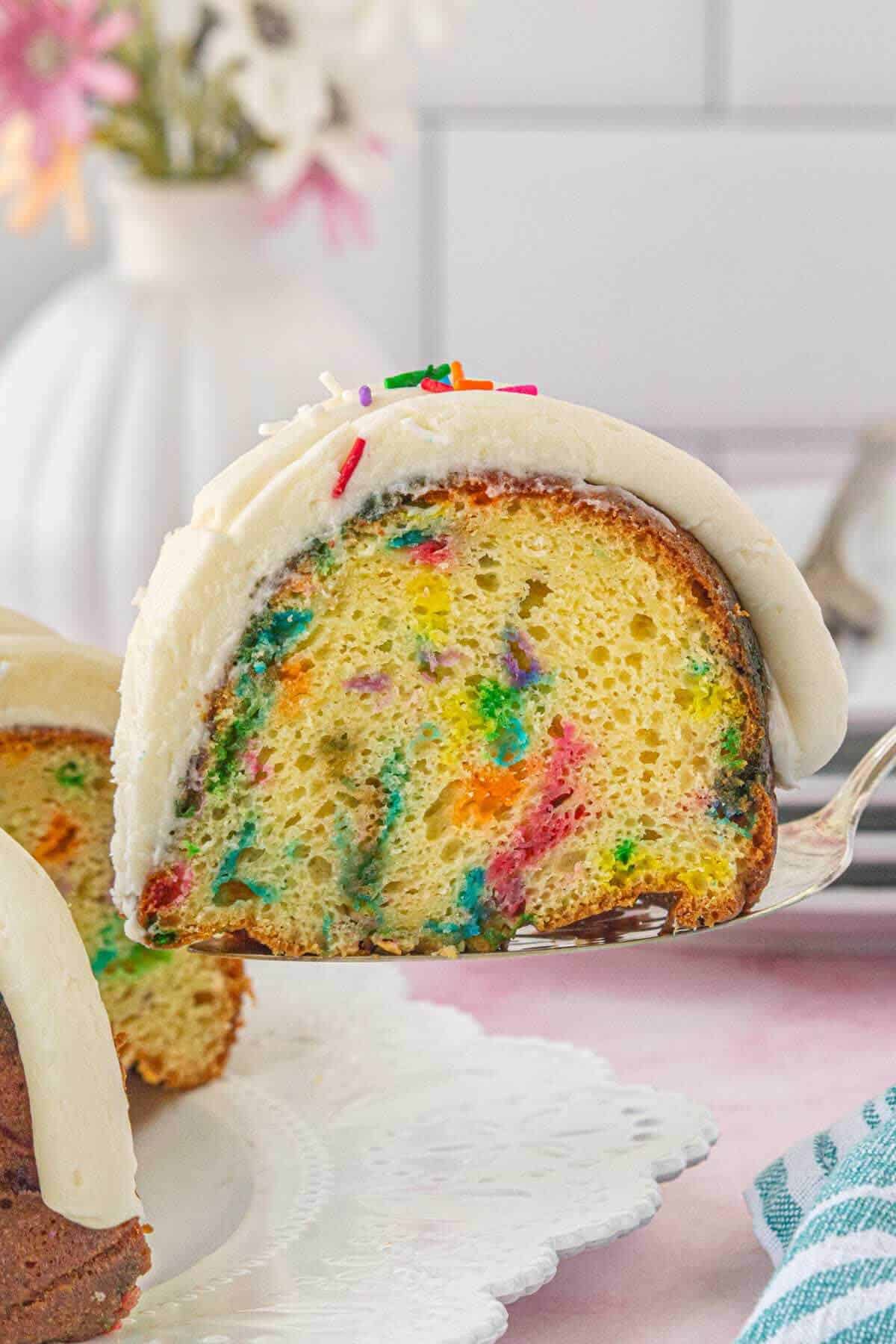 A slice of confetti bundt cake being lifted from the whole cake, showing the bright sprinkle-filled interior and creamy frosting.