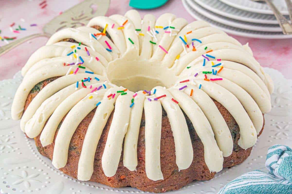 Full bundt cake on a white decorative plate with piped white frosting and rainbow sprinkles, with plates and forks in the background.