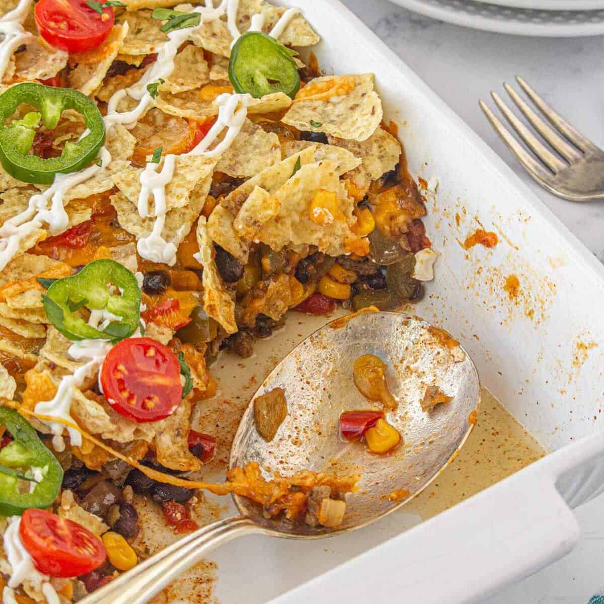 A ground beef casserole in a baking dish, with a serving spoon. The dish is topped with sour cream, tomatoes and sliced jalapenos.