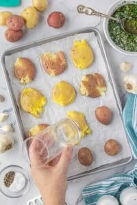 Hand using a glass to gently press and flatten cooked fingerling potatoes on a parchment-lined baking sheet. Surrounding the tray are raw potatoes, garlic, and a bowl of chopped herb mixture.