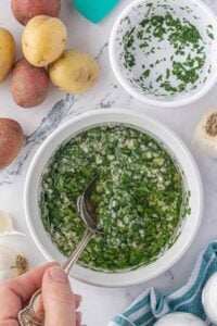 A bowl of fresh garlic herb mixture with olive oil and parsley, being stirred with a silver spoon. Raw potatoes and ingredients are visible nearby.