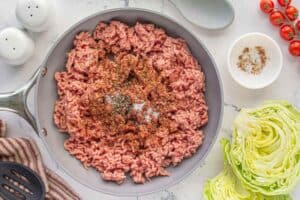 Seasoned ground beef in a skillet surrounded by a head of ice berg lettuce that has been chopped in half.