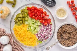 Chopped white onions, cheese, tomatoes and pickles scattered over a layer of iceberg lettuce in a clear mixing bowl.